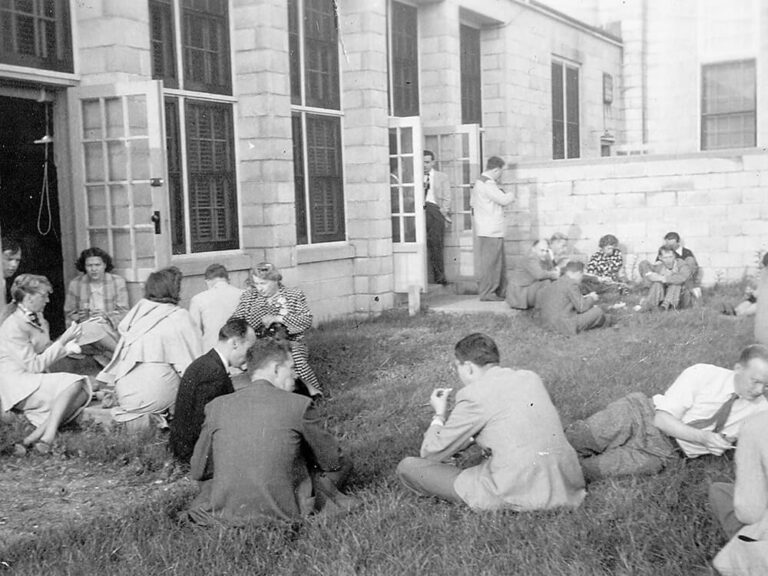 black and white photograph of men and women picnicking on the grass next to a building