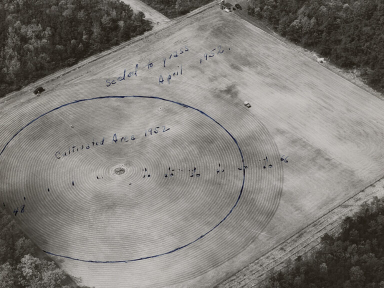 black and white aerial photograph of a field with concentric circular furrows, surrounded by woods