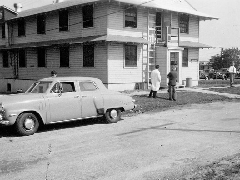 black and white photo of two story wooden building with a 1950s car parked in front